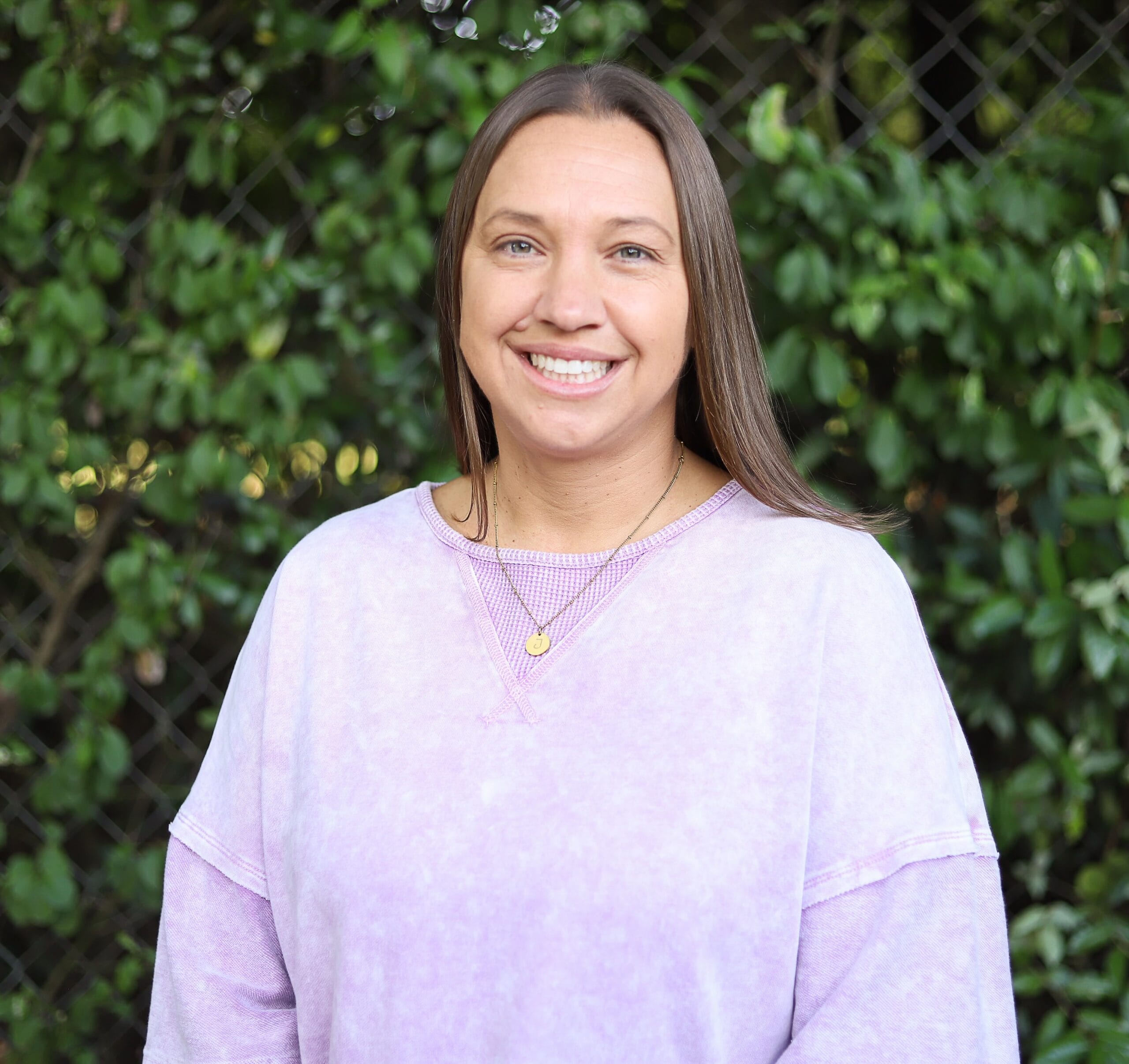Person wearing a lavender shirt smiles in front of lush greenery.