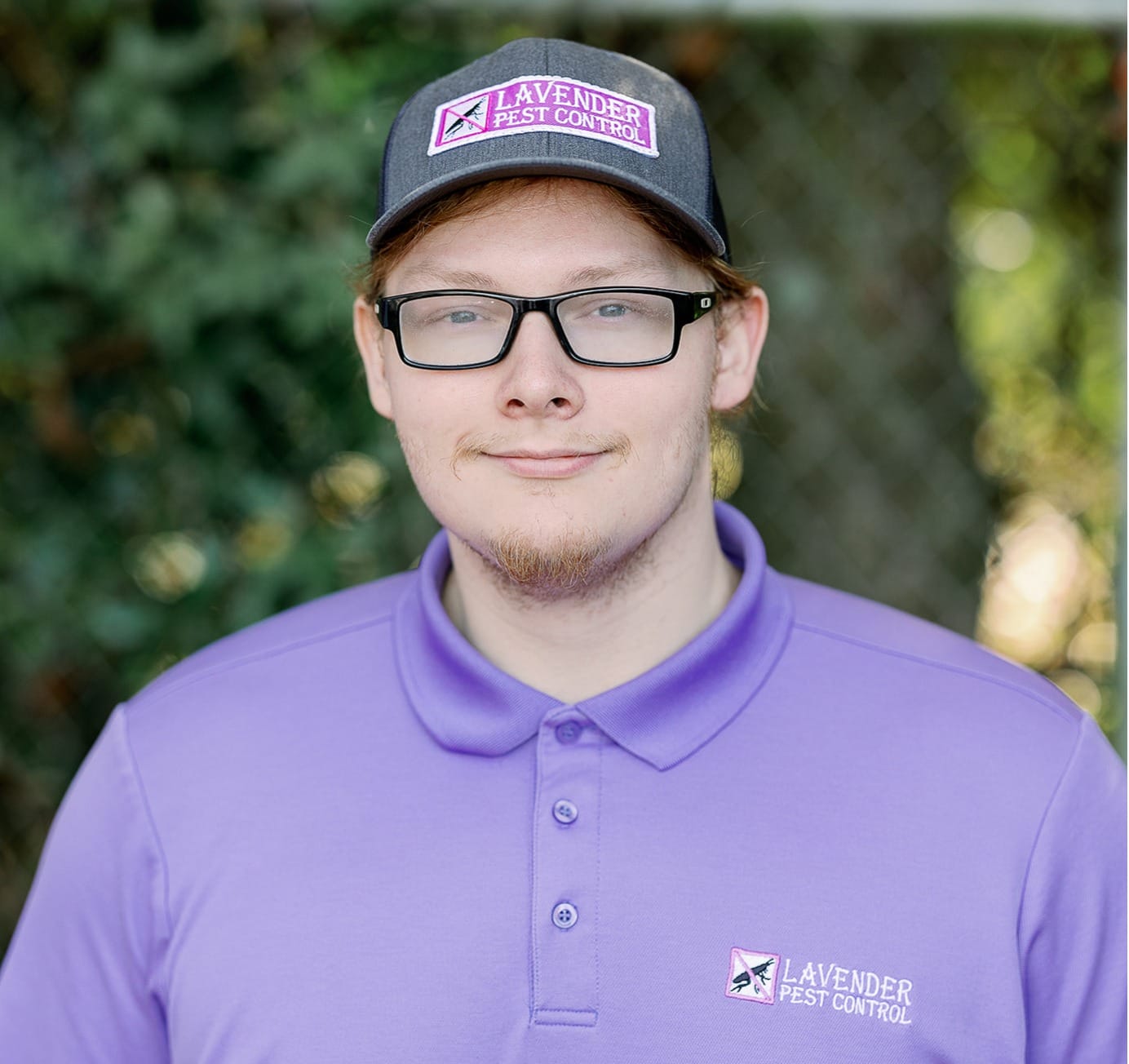Person in a Lavender Pest Control uniform with glasses and cap, standing outdoors.