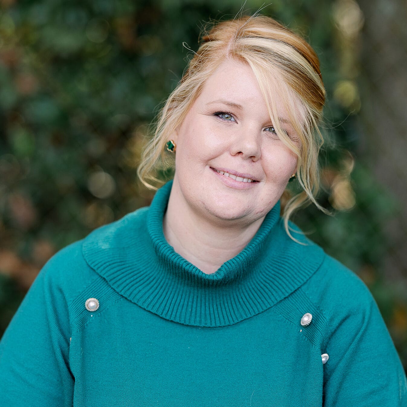 Smiling person in a teal sweater against a blurred foliage background.