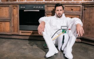 Exhausted pest control worker in protective gear sitting on a kitchen floor, leaning against wooden cabinets.