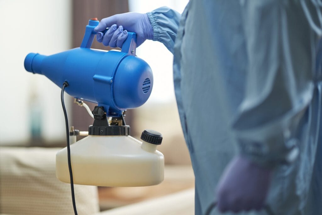 A technician in protective gear holding a blue pest control fogger in a residential setting.